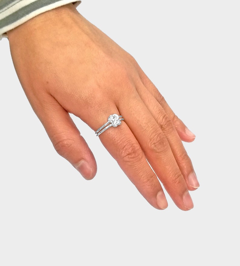 May include: A silver ring with a knot design. The ring is worn on the index finger of a hand.