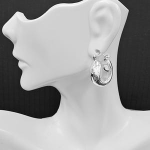 May include: A single, shiny silver hoop earring is displayed on a white mannequin head. The earring is oval-shaped and has a smooth, reflective surface. The earring is attached to the mannequin's ear, showcasing its design and size.