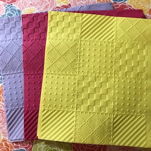 May include: Three square napkins with different embossed patterns. The napkins are purple, red, and yellow.