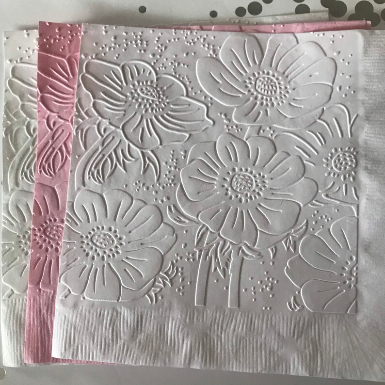 May include: White and pink paper napkins with embossed floral designs. The napkins feature a repeating pattern of large flowers with detailed petals and centers. The edges of the napkins are textured with vertical lines.