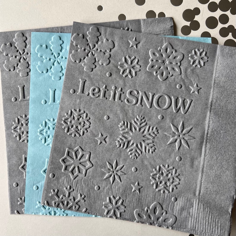 Puede incluir: Tres servilletas de papel grises con un dise&ntilde;o de copo de nieve y el texto "Let it Snow". Las servilletas est&aacute;n apiladas una encima de la otra.