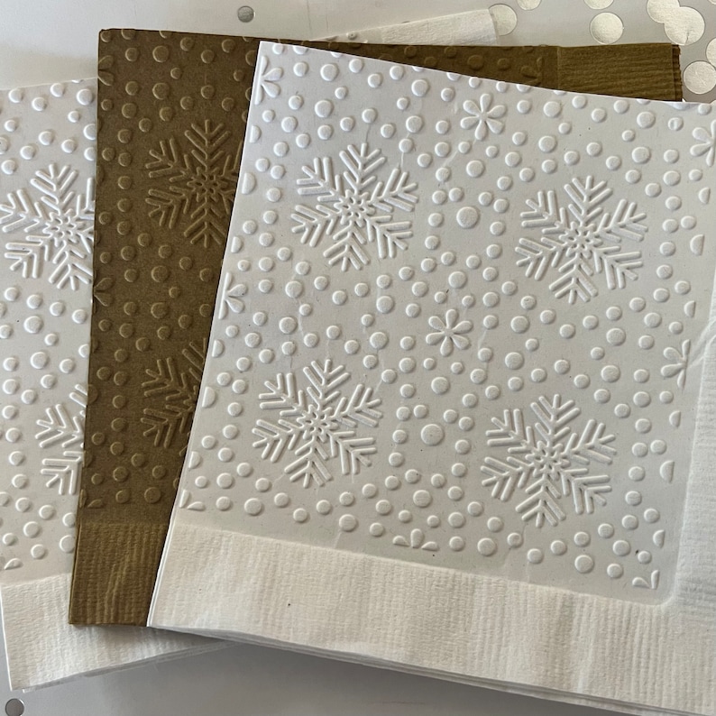 Puede incluir: Una pila de servilletas de papel decorativas con dise&ntilde;os en relieve de copos de nieve y puntos. Las servilletas son blancas y marrones, con un borde blanco en la parte inferior. El dise&ntilde;o en relieve a&ntilde;ade un aspecto texturizado y festivo.