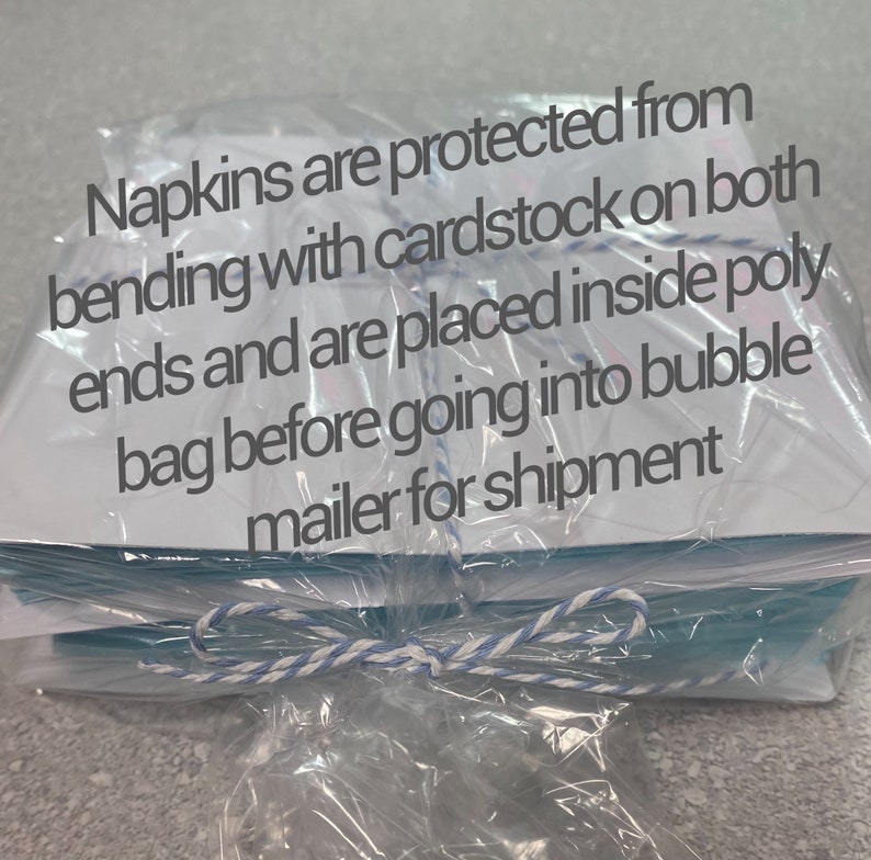 May include: A stack of white napkins, protected with cardstock and wrapped in clear plastic. The napkins are tied with a blue and white string. Text on the plastic reads: "Napkins are protected from bending..."