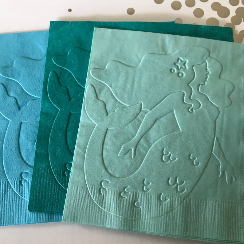May include: Three paper napkins with a mermaid silhouette design. The napkins are in shades of blue and green.