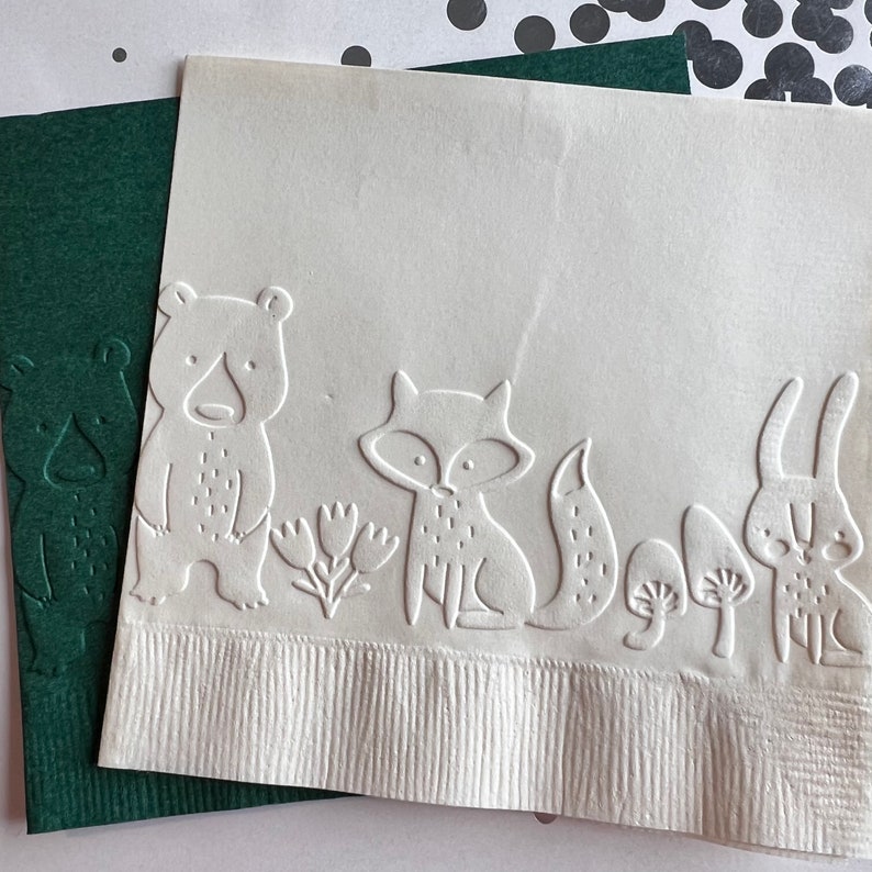 May include: White paper napkins with embossed woodland animal designs, including a bear, fox, and rabbit. The napkins have a textured surface and are placed on a dark green paper. Black and white confetti is in the background.