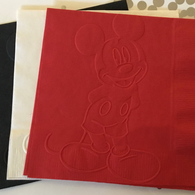 May include: Red paper napkin with an embossed image of Mickey Mouse. Mickey Mouse is smiling and has his signature white gloves on.