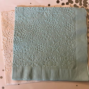 May include: Two paper napkins with embossed snowflake designs. One napkin is light blue, the other is off-white. The snowflakes are of varying sizes and shapes. The napkins are likely for a winter holiday or seasonal event.