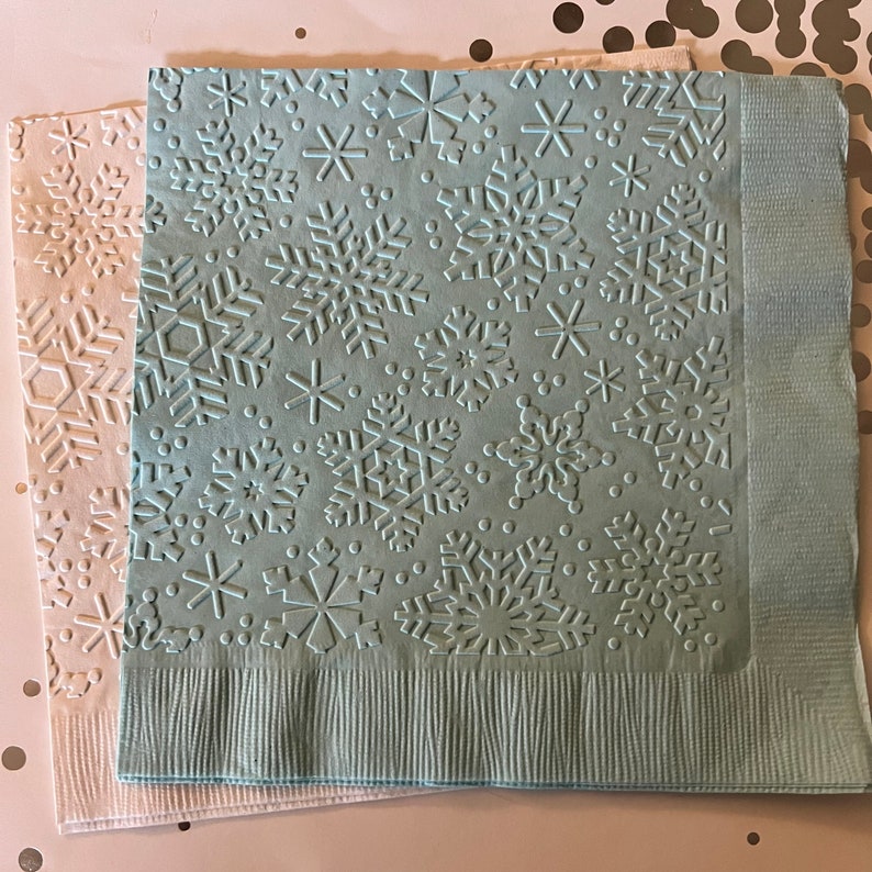 May include: Two paper napkins with embossed snowflake designs. One napkin is light blue, the other is off-white. The snowflakes are of varying sizes and shapes. The napkins are likely for a winter holiday or seasonal event.