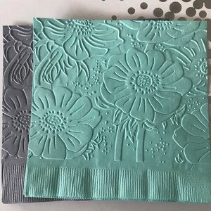 May include: Two decorative paper napkins. One is gray, the other is a light teal. Both have an embossed floral pattern of large flowers and leaves. The bottom edges of the napkins have a fringed border.