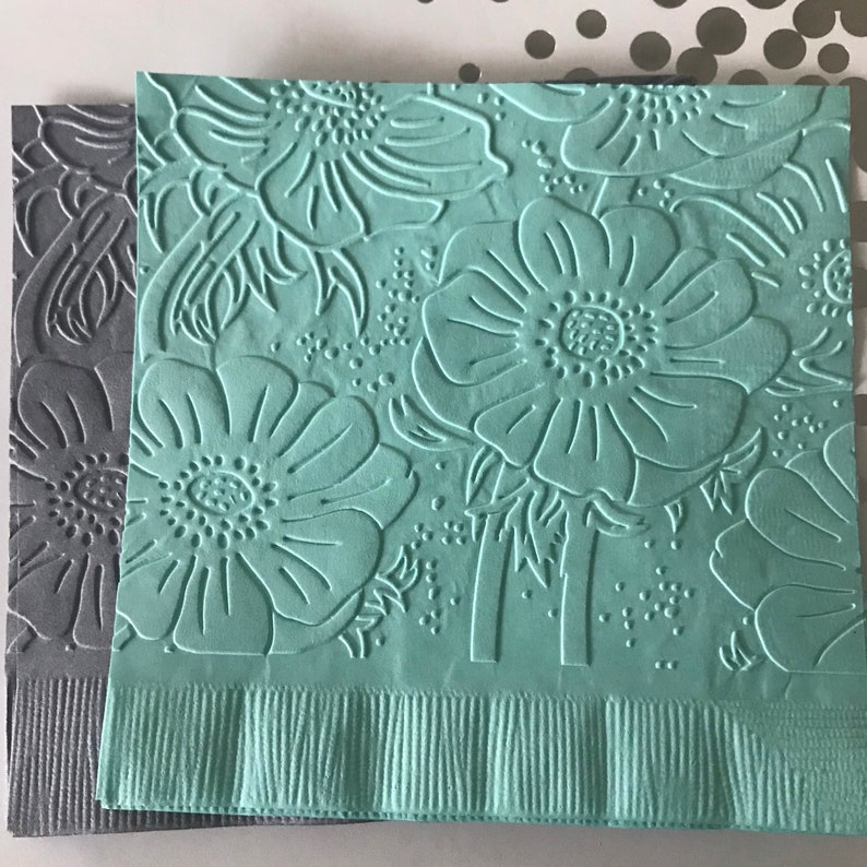 May include: Two decorative paper napkins. One is gray, the other is a light teal. Both have an embossed floral pattern of large flowers and leaves. The bottom edges of the napkins have a fringed border.