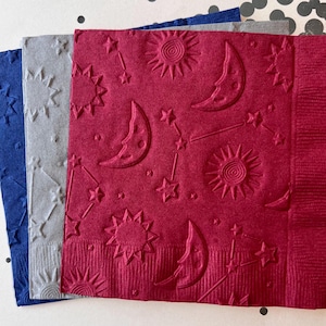May include: Three paper napkins with an embossed design of stars, moons, and suns. The napkins are red, gray, and blue.