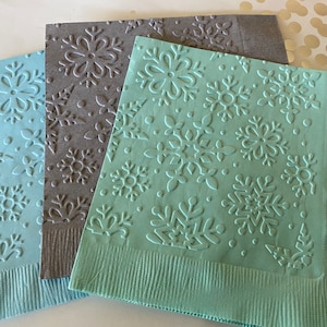 Puede incluir: Primer plano de tres servilletas de papel en relieve en tonos azules y grises. Cada servilleta presenta un patr&oacute;n de copo de nieve. Las servilletas son una adici&oacute;n festiva para las reuniones de invierno.