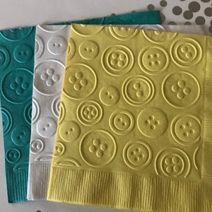 May include: Three paper napkins with a button pattern. The napkins are teal, white, and yellow.