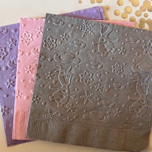 May include: Three paper napkins with a winter theme. The napkins are gray, pink, and purple with a snowflake and snowman design.