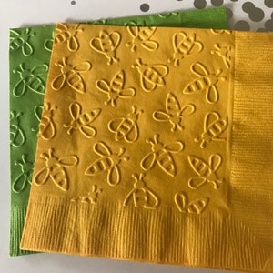 Puede incluir: Dos servilletas de papel amarillas y verdes con un patrón repetido de abejas en relieve. Las servilletas están dobladas por la mitad y apiladas una encima de la otra.