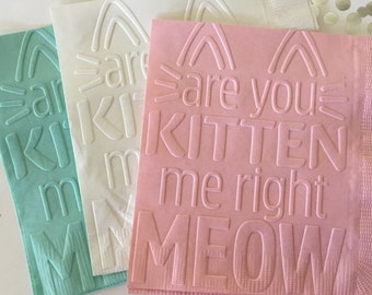 May include: Three paper napkins with a raised, white, embossed design. The napkins are in three colors: light blue, white, and pink. The pink napkin reads "are you kitten me right meow".