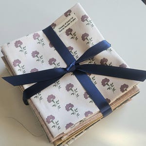 May include: A stack of fabric squares tied with a navy blue ribbon. The fabric has a cream background with a repeating pattern of purple flowers and green leaves. The label reads "Threads of Grace Fat Quarter Bundle 14 piece."