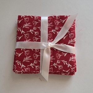 May include: A stack of red fabric squares with a white floral pattern, tied with a white satin ribbon. The fabric is a deep red with a delicate white outline of flowers and leaves. The ribbon is tied in a bow.