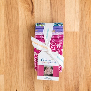 May include: A stack of fabric strips in shades of pink, purple, and teal, tied with a white ribbon. A tag attached to the ribbon features the text Chasing Rainbows, a photograph of a person, and the Riley Blake Designs logo in blue and pink.