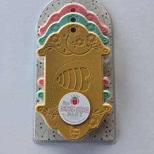 May include: A set of three Bee Binding Holders in a clear plastic package. The holders are in shades of pink, green, and gold. The gold holder has a floral design and the text "Bee Binding Holder Set of 3".