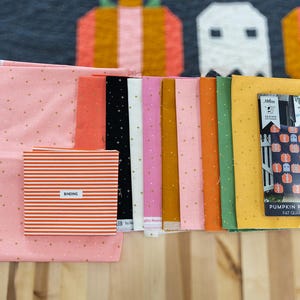 May include: A fabric bundle for a quilt project featuring various solid and star-patterned cotton squares in shades of pink, black, white, orange, yellow, and green. Includes a striped fabric labeled BINDING and a Pumpkin Patch Ghosts pattern.