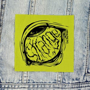 May include: A square lime green fabric patch featuring a black graphic illustration of a stylized eye with a textured iris. The text Lofty Dog Designs is printed vertically in black on the right side of the eye graphic.