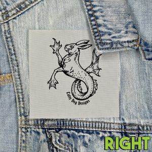 May include: A light gray patch featuring a black illustration of a mythical creature with a rabbit head and fish tail. The text "Lofty Dog Designs" is below the creature. The patch is on a denim jacket.