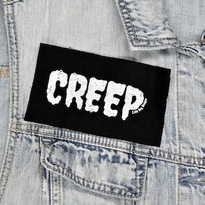 Op de afbeelding: Een zwart rechthoekig embleem met het woord "CREEP" in verweerde witte letters. De patch is bevestigd aan een lichtblauwe spijkerjas. De tekst "Lofty Dog Designs" is in het wit gedrukt in de rechter benedenhoek.