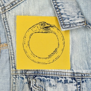 May include: A yellow square patch featuring a black ouroboros design, a snake eating its own tail. The patch is sewn onto a light blue denim jacket. The text "Lofty Dog Designs" is printed on the patch.