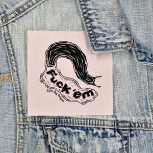 Puede incluir: Un parche rosa con las palabras "Fuck 'em" en negro, junto con un diseño gráfico negro. El parche está cosido a una chaqueta vaquera azul claro. El texto "Lolly Dog Designs" está impreso debajo.