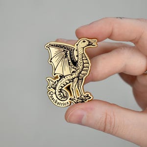 Pode incluir: Pin de metal dourado com uma ilustração em preto e branco de uma criatura mítica com cabeça de pássaro, asas e corpo de serpente. O texto "Quackatrice" está impresso abaixo da criatura.