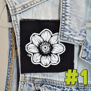 May include: Black and white flower patch with the letters "LDD" on the lower left corner. The patch is on a black background and is attached to a denim jacket. The number "#1" is in green on the lower right corner.