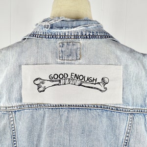 Könnte beinhalten: Eine hellblaue Jeansjacke mit einem weißen Patch, der auf der Rückseite aufgenäht ist. Der Patch zeigt eine Schwarzweißillustration eines gebrochenen Knochens mit Bandagen und dem Text "GOOD ENOUGH".