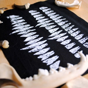 May include: A black fabric with a white print of rows of teeth. The fabric is surrounded by various animal bones and claws.