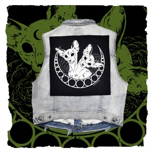 Peut inclure: Un gilet en jean noir avec un patch blanc représentant deux chats stylisés avec trois yeux chacun, entourés d'un croissant de lune et d'un cercle de petits cercles. Le texte "Love the Strange" est en bas du patch.