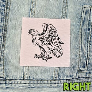 Op de afbeelding: Een vierkant, gebroken wit embleem met een zwarte illustratie van een vogel met visachtige kenmerken en uitgespreide vleugels. De patch is genaaid op een lichtblauwe spijkerjas. De tekst "Caty Bug Designs" is op de patch gedrukt.