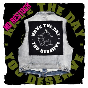 May include: A light blue denim vest featuring a large square black patch on the back. The patch displays a white graphic of a fist with a bone protruding from the wrist, surrounded by the text HAVE THE DAY YOU DESERVE in white capital letters.