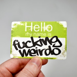 May include: A rectangular sticker with a green and white background. The sticker reads "Hello my name is fucking weirdo" in black, bold lettering. The sticker is held by a hand.