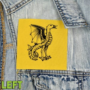 May include: A yellow square patch with a black illustration of a dragon-like creature with a duck's head and wings. The patch is sewn onto a denim jacket. The text "Lofty Dog Designs" is printed on the patch.