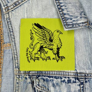 Może przedstawiać: Kwadratowa, limonkowo-zielona naszywka z czarnym wzorem gryfa. Gryf, mityczne stworzenie, ma ciało lwa oraz głowę i skrzydła orła. Tekst "Lofty Dog Designs" jest wydrukowany po lewej stronie naszywki.