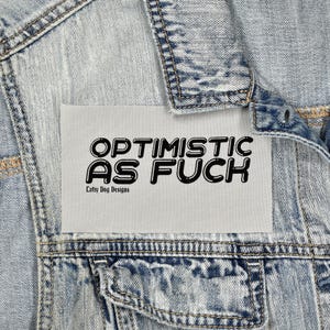 May include: A light blue denim jacket with a rectangular patch. The patch features the text "OPTIMISTIC AS FUCK" in bold, black, bubble letters. The text "Lofty Dog Designs" is below the main text.