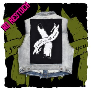 May include: A light blue denim vest featuring a large rectangular black patch on the back. The patch displays a white graphic of a tattered ribbon crossed over itself with the text I wish you the worst printed in a cursive font across the center.