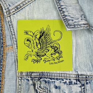 May include: A square, lime green patch featuring a black illustration of a three-headed mythical creature with wings and a curled tail. The text "Lofty Dog Designs" is printed in black on the bottom right corner. The patch is sewn onto a denim jacket.