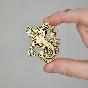 May include: Gold-colored metal pin featuring a black and white illustration of a hippocampus, a mythical creature with the head and forelegs of a horse and the tail of a fish. The pin has the word "Hippocampus" inscribed on the bottom.