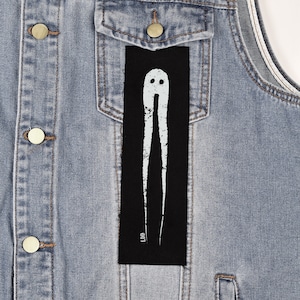 Op de afbeelding: Een zwart rechthoekig embleem met een wit, abstract spookachtig ontwerp. Het embleem is bevestigd aan een lichtblauwe spijkerjas. Het ontwerp heeft twee verticale lijnen en een afgeronde bovenkant met twee kleine cirkels. De letters "LDD" staan onderaan.