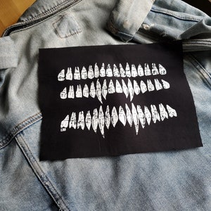 May include: A black patch with a white design of three rows of teeth. The patch is sewn onto a light blue denim jacket.