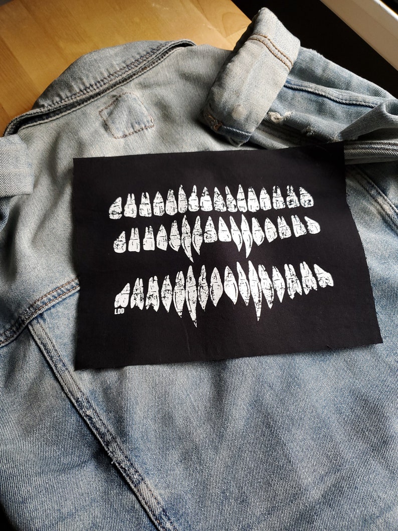 May include: A black patch with a white design of three rows of teeth. The patch is sewn onto a light blue denim jacket.