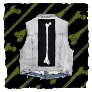 May include: A light-wash denim vest featuring a long, rectangular black patch centered on the back. The patch displays a detailed white line-art illustration of a human femur bone, extending vertically from the top to the bottom of the fabric.