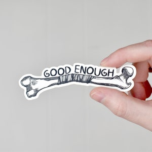以下が含まれることがあります： 包帯で包まれた折れた骨の白黒のイラストが描かれた白いステッカー。骨の上に「GOOD ENOUGH」という文字が印刷されています。