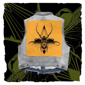 May include: A light blue denim vest featuring a large, square, mustard-yellow patch on the back. The patch displays a detailed black ink illustration of a stag beetle with an eye in its center, positioned over a geometric triangle design.
